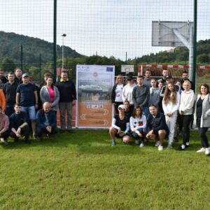 Studenci i wykładowcy PANS na tle rollupu projektowego na boisku sportowym podczas wycieczki turystyczno-krajoznawcza śladami dawnej galicyjskiej linii kolejowej z Przemyśla do Budapesztu