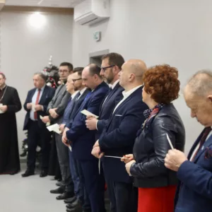 goście stojący w sali podczas spotkania opłatkowego, w rękach trzymają opłatek