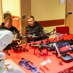 dwóch studentów przy stanowisku z dronami, na stole leżą ulotki, długopisy i smycze PANS