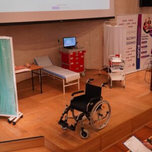scena w auli, po lewej stronie parawan, po prawej mównica, na pierwszym planie wózek inwalidzki, z tyłu łóżko szpitalne