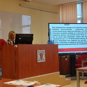 prelegentka przy komputerze, obok tablica multimedialna z prezentacją
